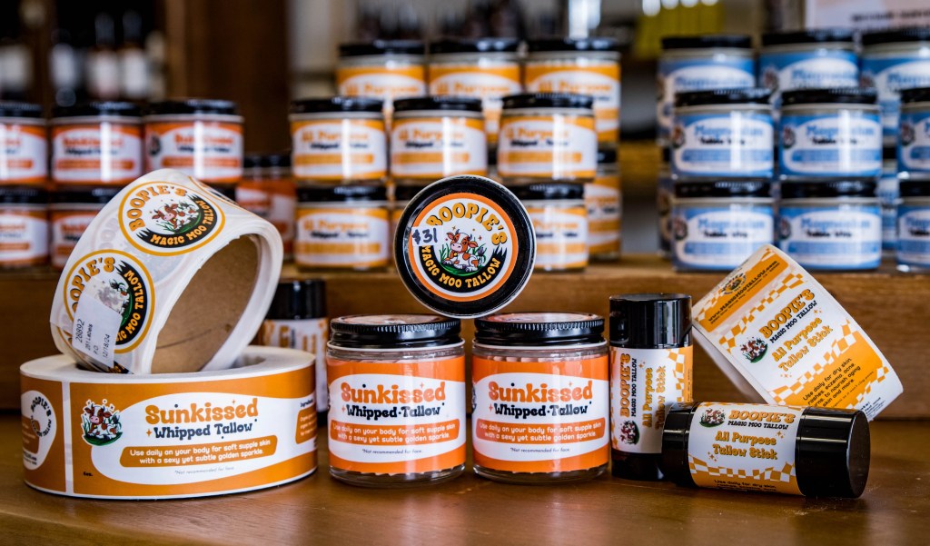 A collection of Magic Moo Tallow products, featuring small jars of tallow and Tallow Sticks. Each product is accompanied by rolls of labels, which are being applied to the jars and sticks. The jars are neatly arranged, showcasing the branding and the natural appearance of the tallow.